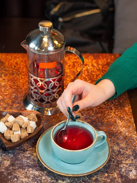 Fruit red tea is poured into a mug. A woman\'s hand is stirring sugar with a spoon. Next to it is a chaikik fren press and a plate of white and cane sugar. Vitamin teas.