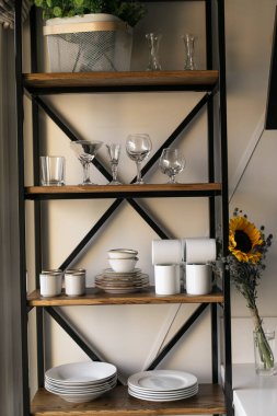 A rack of white dishes stands against a white wall. Next to it is a bouquet of wildflowers: sunflowers and blue thorns. The kitchen is in the style of Provence. Summer mood. Shelf in the kitchen. 
