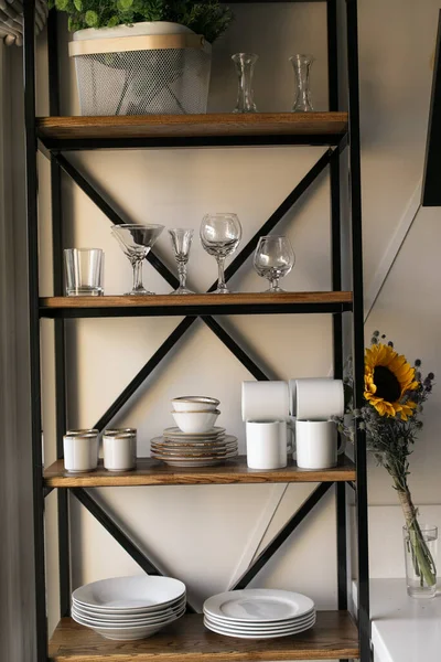 A rack of white dishes stands against a white wall. Next to it is a bouquet of wildflowers: sunflowers and blue thorns. The kitchen is in the style of Provence. Summer mood. Shelf in the kitchen. 