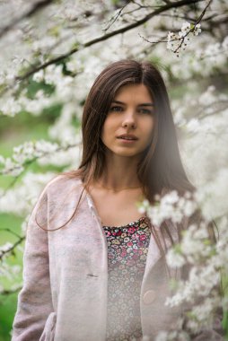 Kiraz ağacında güzel bir kız. Genç Avrupalı esmer kadın portresi doğal arka planda, bahar mevsimi, çiçek açan bahçe. Bahar pastel renginde giysiler giyiyor.