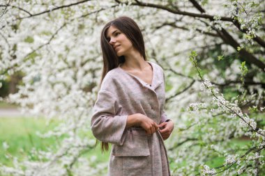 Pastel renkli ceketli, kiraz ağacıyla güzel bir kız. Genç Avrupalı esmer kadın portresi doğal arka planda, bahar mevsimi, çiçek açan bahçe. Bahar kıyafetleri giyiyor.