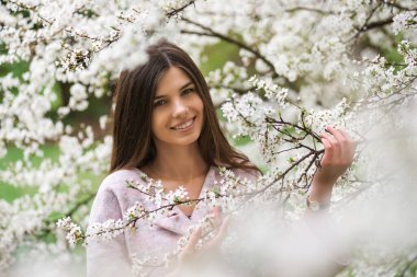 Çiçekli kiraz ağacında gülümseyen güzel bir kız. Genç Avrupalı esmer kadın portresi doğal arka planda, bahar mevsimi, çiçek açan bahçe. Bahar pastel renginde giysiler giyiyor.