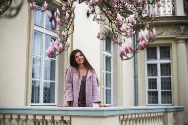 Pastel ceketli ve terasta çiçekli elbisesi olan hoş bir kız. Pembe çiçekli manolya ağacının altında. Mimari arka planda genç Avrupalı esmer kadın portresi, bahar mevsimi, çiçek açıyor. Bahar kıyafetleri giyiyor.