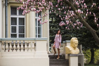 Pastel ceketli ve çiçekli elbiseli kız, terasta pembe çiçekli manolya ağacının altında kalıyor. Mimari arka planda genç Avrupalı esmer kadın portresi, bahar mevsimi, çiçek açıyor. Bahar kıyafetleri giyiyor.