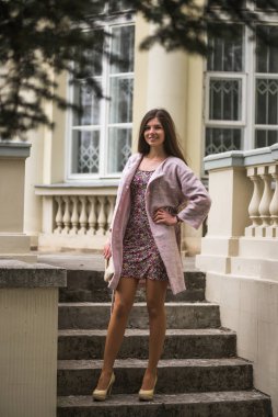 Pastel ceketli, çiçekli, pembe çiçekli manolya ağacıyla güzel bir kız. Genç Avrupalı esmer kadın portresi mimari arka planda, bahar mevsimi, çiçek açan teras. Bahar kıyafetleri giyiyor.