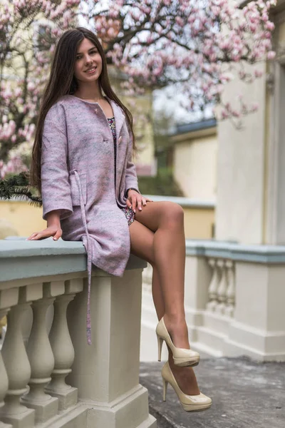 Kremalı pastel ceketli, çiçekli elbisesi olan, pembe çiçekli manolya ağacı olan güzel bir kız. Genç Avrupalı esmer kadın portresi mimari arka planda, bahar mevsimi, çiçek açan teras. Bahar kıyafetleri giyiyor.