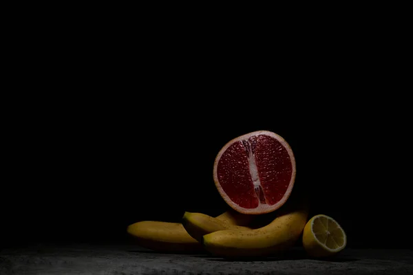 A juicy red grapefruit rests on three bananas, next to a lemon on gray concrete. Low key.
