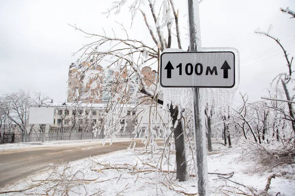 An icy road sign 100 meters in winter showing the direction of travel against a background of icy road and trees.