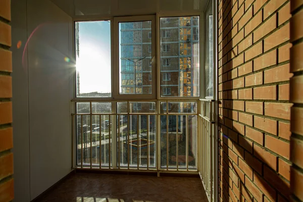 El sol brillante brilla maravillosamente a través de la ventana en el balcón con barras y ventanas de plástico. Echa una sombra sobre una pared de ladrillo. Imagen De Stock