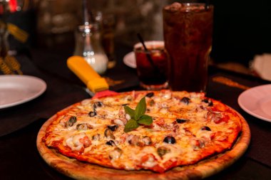 Masadaki ahşap tabakta deniz ürünü ve peynirli iştah açıcı bir pizza yatıyor. Soğuk, kahverengi bir içecekle.