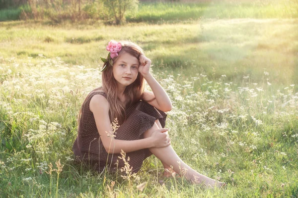 Девушка с пионом в волосах сидит на поле и смотрит — стоковое фото