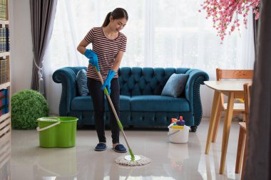 Genç Asyalı mutlu ev hanımı eldiven giyip oturma odasında yerleri paspasla yıkıyor, temizlik yapıyor.
