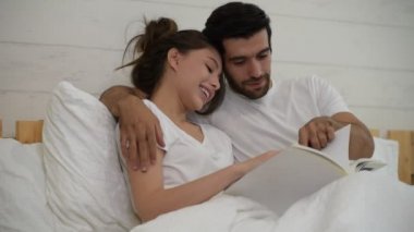 Mutlu, genç, rahat, sevgi dolu bir çift yatakta uzanıp evde birlikte kitap okuyor.