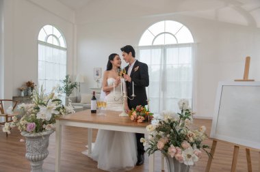 Romantik Asyalı gelin ve damat yakın durup, samimi bir anda gülümsüyor ve burunlarına dokunuyorlar. Damat siyah bir takım giyer ve gelin beyaz bir gelinlik giyer. Zarif düğün sahnesi.