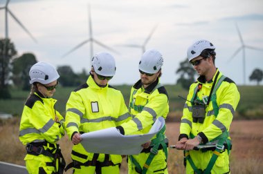 Güvenlik kaskı takan mühendisler, rüzgar tarlasında çalışırken rüzgar türbinini inceliyorlar. Yenilenebilir enerji sürdürülebilir teknoloji rüzgar enerjisi bakım çevre mühendisliği.