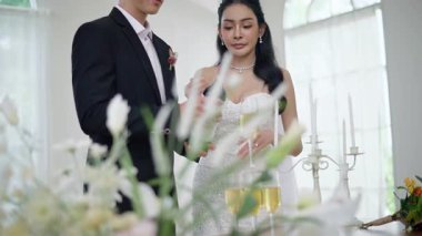 Düğün kıyafetleri içinde mutlu bir Asyalı çift, gelin güzel beyaz bir gelinlik ve damatla klasik bir takım elbise içinde düğün resepsiyonunda şampanya içiyor.