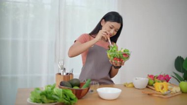 Önlük takan genç bir Asyalı kadın mutfakta sağlıklı malzemeler ve sebzelerle yemek için taze ev yapımı salata yapıyor. Sağlıklı refah kavramı.