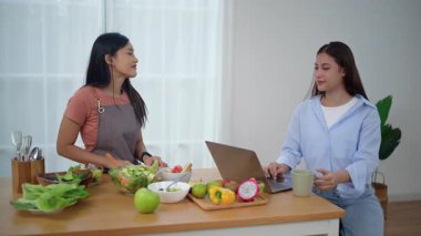 Önlük takan genç bir Asyalı kadın evde, mutfakta sağlıklı malzemeler ve sebzelerle durup arkadaşlarıyla tatilde çalışan arkadaşlarıyla birlikte yemek için taze ev yapımı salata yapıyor. Sağlıklı konsept.