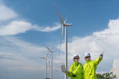 Rüzgâr tarlasında rüzgar türbinini denetleyen ve çalışırken dizüstü bilgisayar tutan güvenlik kaskı takan mühendisler. Yenilenebilir enerji sürdürülebilir teknoloji rüzgar enerjisi bakım çevre mühendisliği.