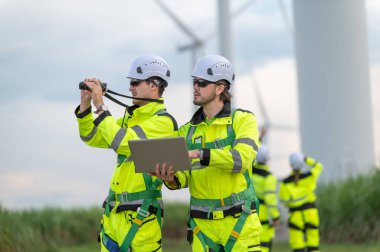 Rüzgâr tarlasında rüzgar türbinini denetleyen ve çalışırken dizüstü bilgisayar tutan güvenlik kaskı takan mühendisler. Yenilenebilir enerji sürdürülebilir teknoloji rüzgar enerjisi bakım çevre mühendisliği.
