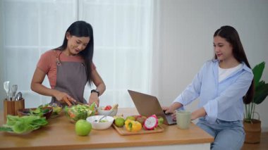 Önlük takan genç bir Asyalı kadın evde, mutfakta sağlıklı malzemeler ve sebzelerle durup arkadaşlarıyla tatilde çalışan arkadaşlarıyla birlikte yemek için taze ev yapımı salata yapıyor. Sağlıklı konsept.