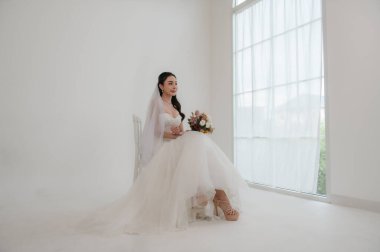 Parlak bir stüdyoda çiçeklerle çevrili bir koltukta otururken beyaz gelinlik içinde, gülümseyen ve yüzüğüne hayran olan zarif bir gelin..