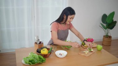Önlük takan genç bir Asyalı kadın mutfakta sağlıklı malzemeler ve sebzelerle yemek için taze ev yapımı salata yapıyor. Sağlıklı refah kavramı.