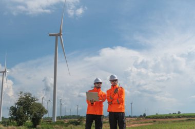 Rüzgâr tarlasında rüzgar türbinini denetleyen ve çalışırken dizüstü bilgisayar tutan güvenlik kaskı takan mühendisler. Yenilenebilir enerji sürdürülebilir teknoloji rüzgar enerjisi bakım çevre mühendisliği.