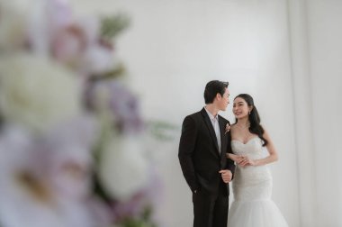 Düğün kıyafetleri içinde mutlu Asyalı gelin ve damat gülümseyip birbirlerine bakıyorlar, evin içinde parlak pencere ışığında duruyorlar ve ön planda çiçek süslemeleri var. Düğün için romantik çift portresi.