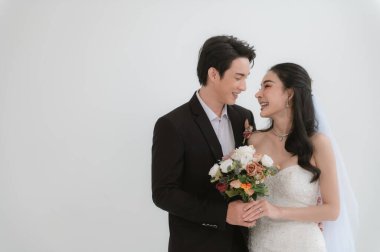 Asyalı çift evleniyor, mutlu Asyalı çift düğün kıyafetleri içinde, gelin güzel beyaz bir gelinlik ve damatla klasik bir takım elbise giyiyor, ikisi de çekici ve aşık görünüyorlar.