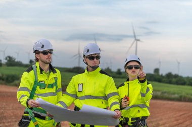 Güvenlik kaskı takan mühendisler, rüzgar tarlasında çalışırken rüzgar türbinini inceliyorlar. Yenilenebilir enerji sürdürülebilir teknoloji rüzgar enerjisi bakım çevre mühendisliği.
