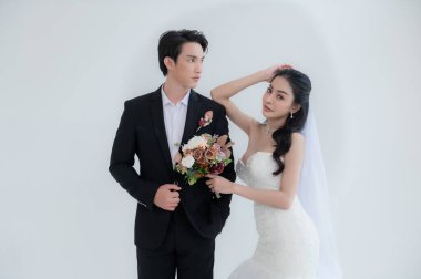 Asyalı çift evleniyor, mutlu Asyalı çift düğün kıyafetleri içinde, gelin güzel beyaz bir gelinlik ve damatla klasik bir takım elbise giyiyor, ikisi de çekici ve aşık görünüyorlar.