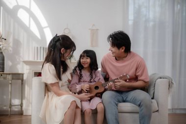 Mutlu Asyalı aile kızına evde ukulele çalmayı öğretiyor, ebeveynler ve çocuklar rahat oturma odasında müzik dinlemeyi, müzik eğitimini, ebeveynliği, aşkı, evde öğrenmeyi ve sıcak aile yaşamını seviyorlar..