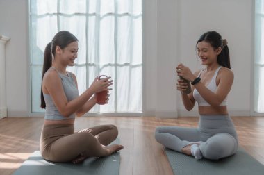 Spor kıyafetleri giyen genç kadınlar, su şişeleriyle antreman yaptıktan sonra dinleniyorlar. Oturma odasında egzersiz yaptıktan sonra gülümsüyorlar. Spor ve dinlenme konsepti..