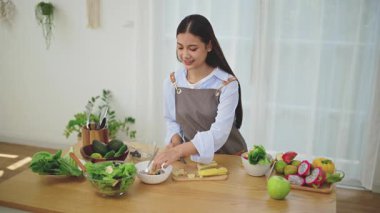 Önlük takan genç bir Asyalı kadın mutfakta sağlıklı malzemeler ve sebzelerle yemek için taze ev yapımı salata yapıyor. Sağlıklı refah kavramı.