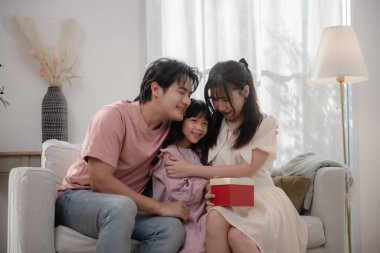 Mutlu Asyalı aile evdeki hediye kutusunu açarken birlikte kanepede oturuyor. Baba, anne ve kız oturma odasında neşeli bir sürpriz anı paylaşıyorlar. Aile sevgisi, mutluluk, kutlama.