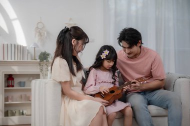 Mutlu Asyalı aile kızına evde ukulele çalmayı öğretiyor, ebeveynler ve çocuklar rahat oturma odasında müzik dinlemeyi, müzik eğitimini, ebeveynliği, aşkı, evde öğrenmeyi ve sıcak aile yaşamını seviyorlar..