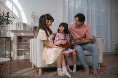 Mutlu Asyalı aile kızına evde ukulele çalmayı öğretiyor, ebeveynler ve çocuklar rahat oturma odasında müzik dinlemeyi, müzik eğitimini, ebeveynliği, aşkı, evde öğrenmeyi ve sıcak aile yaşamını seviyorlar..