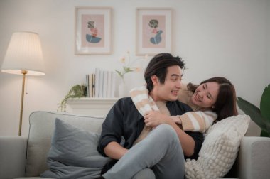 Mutlu genç Asyalı çift sarılıyor, rahat bir kanepede birlikte oturuyor, aşırı mutlu bir şekilde gülen kadın ve erkek eğleniyor, boş vakitlerin tadını çıkarıyor, evdeki oturma odasında kanepede dinleniyor, iyi bir ilişki içinde.