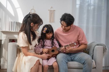 Mutlu Asyalı aile kızına evde ukulele çalmayı öğretiyor, ebeveynler ve çocuklar rahat oturma odasında müzik dinlemeyi, müzik eğitimini, ebeveynliği, aşkı, evde öğrenmeyi ve sıcak aile yaşamını seviyorlar..