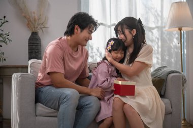 Asyalı ailelerin evdeki kanepede kızlarına hediye kutusuyla sarılmaları. Aile sevgisi, ebeveynlik, kutlama, minnettarlık, sürpriz hediye, aşk ve mutlu ev hayatı kavramı..