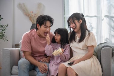 Mutlu Asyalı aile sinemada birlikte eğleniyor, ebeveynler ve kızları kanepelerde atıştırmalıkları paylaşıyor, aile bağları, sevgi, mutluluk, boş zaman ve sıcak ev hayatı kavramı.