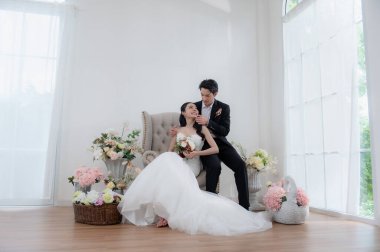 Mutlu Asyalı gelin ve damat, temiz bir arka planla içeride poz veriyorlar. Düğün fotoğrafı konsepti için şık bir damat portresi. İkisi de çekici ve aşık görünüyor..