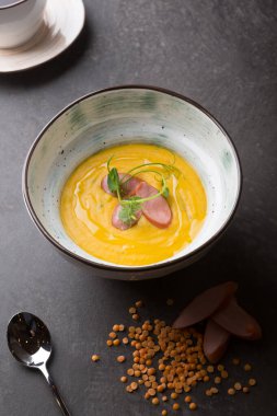 Sarı mercimek kremalı çorba, siyah bir masada sosis ile servis edilir.