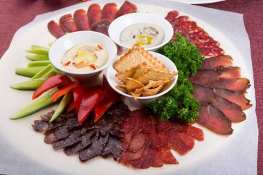 Humus ve sebzelerle servis edilen çeşitli dilimlenmiş tuzlu ve kurutulmuş et tabağı.