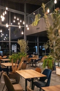 Geceleri modern bir restoranın içinde ahşap masalar, karışık bej ve mavi sandalyeler, yerden tavana geniş pencereler, dekoratif ışıklar ve kapalı mekandaki bitkiler yer alıyor. Konukseverlikle ilgili konular için çağdaş yemek alanı, di