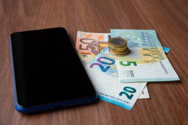 Yanında paralar ve banknotlar olan ahşap yüzeyde akıllı bir telefon. Telefon faturaları ve ödemeler.