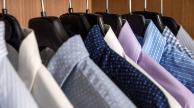 Çeşitli renklerde gömlekler, ahşap renkli bir gardıropta askılarda yan yana asılıydı.