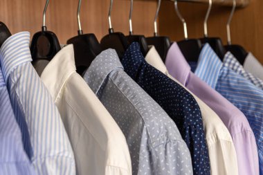 Çeşitli renklerde gömlekler, ahşap renkli bir gardıropta askılarda yan yana asılıydı.