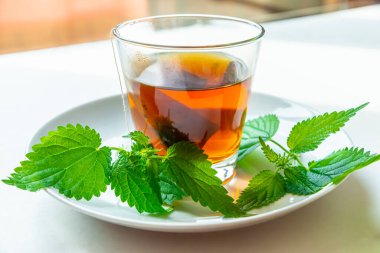 Şeffaf bir fincanda, suya batırılmış bir kese ve yanında ısırgan otu yaprakları olan beyaz bir fincan bitkisel çay. Isırgan otunun iyileşme aşısı. 